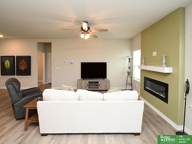 living room with light wood-style flooring, a ceiling fan, a glass covered fireplace, and baseboards