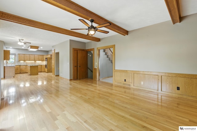 10428 Monroe St, Omaha, NE 68127 - unfurnished living room featuring ceiling fan, beam ceiling, light wood finished floors, stairway, and a wainscoted wall