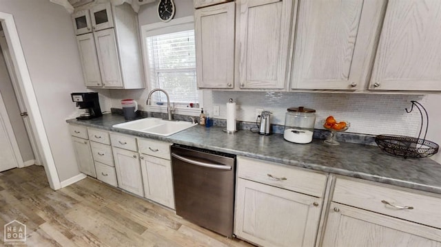 kitchen with light wood-style floors, stainless steel dishwasher, a sink, dark countertops, and backsplash