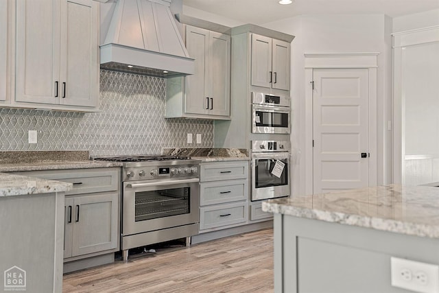 kitchen featuring tasteful backsplash, light wood-type flooring, appliances with stainless steel finishes, premium range hood, and light stone countertops