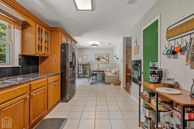 603 Arrowhead Dr, Jonesboro, AR 72401 - kitchen featuring stainless steel refrigerator with ice dispenser, black oven, light tile patterned floors, glass insert cabinets, and brown cabinetry