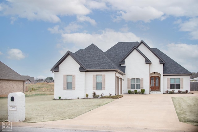 french country inspired facade with driveway, french doors, a front lawn, fence, and a shingled roof