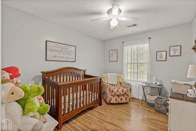 bedroom with a crib, a ceiling fan, wood finished floors, and visible vents