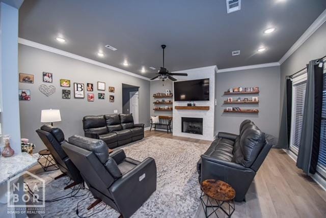 living area with light wood-type flooring, a large fireplace, ornamental molding, a ceiling fan, and visible vents