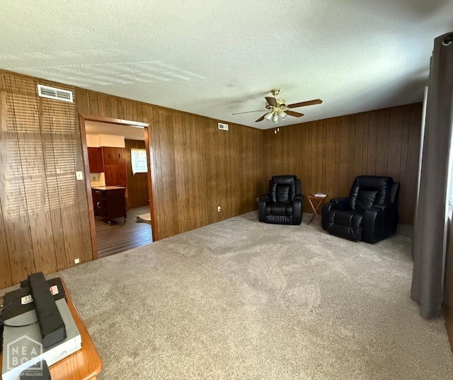 sitting room with wood walls, a textured ceiling, visible vents, carpet floors, and a ceiling fan