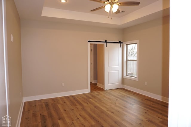spare room with wood finished floors, a ceiling fan, a barn door, baseboards, and a raised ceiling