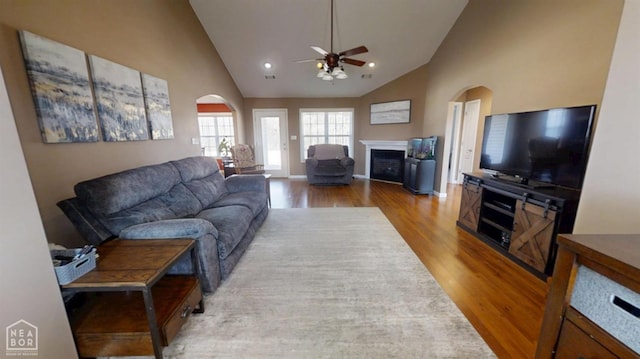 living room featuring a fireplace, a ceiling fan, high vaulted ceiling, arched walkways, and wood finished floors