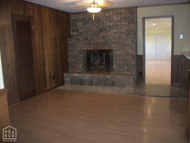 unfurnished living room with a brick fireplace, a ceiling fan, wainscoting, wood finished floors, and wood walls