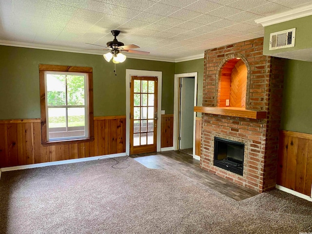 unfurnished living room with crown molding, a brick fireplace, wooden walls, carpet floors, and ceiling fan