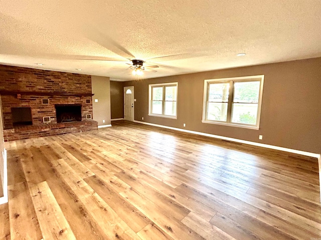 unfurnished living room featuring a fireplace, brick wall, ceiling fan, a textured ceiling, and light hardwood / wood-style flooring