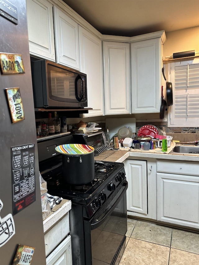 kitchen with black gas range oven, stainless steel fridge, sink, light tile patterned floors, and white cabinets