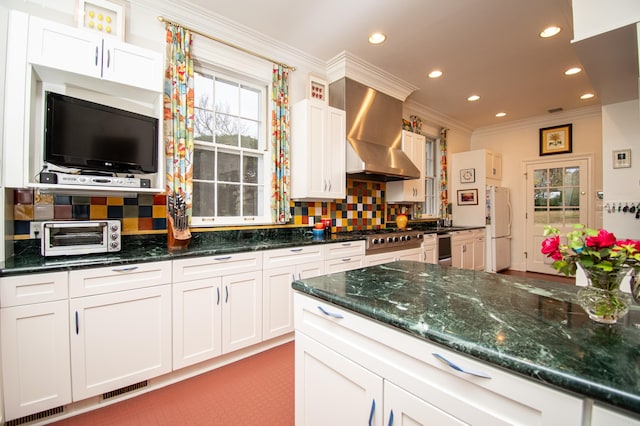 kitchen featuring dark stone counters, white fridge, wall chimney range hood, decorative backsplash, and stainless steel gas stovetop