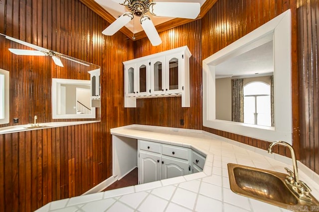 kitchen featuring ceiling fan, sink, white cabinets, wooden walls, and tile counters