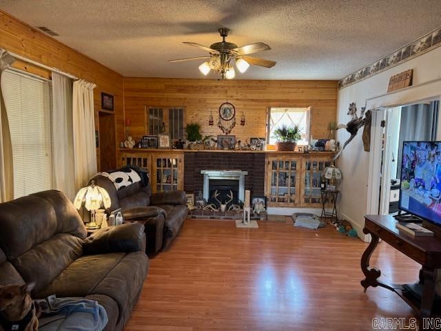 living room featuring ceiling fan, a brick fireplace, a textured ceiling, wood-type flooring, and wood walls