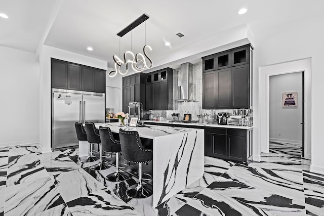 kitchen featuring wall chimney exhaust hood, stainless steel appliances, an island with sink, pendant lighting, and decorative backsplash