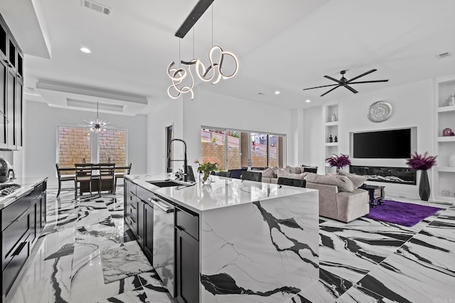 kitchen with a kitchen island with sink, hanging light fixtures, sink, built in shelves, and light stone counters