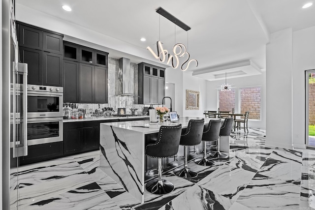 kitchen featuring wall chimney range hood, tasteful backsplash, a notable chandelier, decorative light fixtures, and a kitchen island with sink