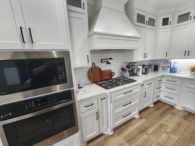 kitchen featuring custom range hood, appliances with stainless steel finishes, tasteful backsplash, light hardwood / wood-style floors, and white cabinetry