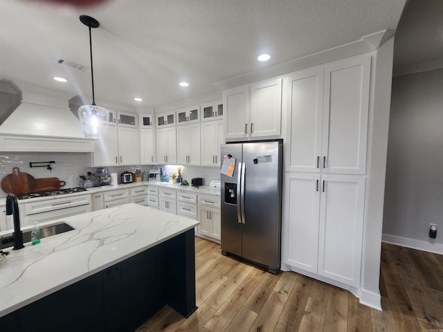 kitchen with light stone countertops, custom exhaust hood, stainless steel appliances, decorative light fixtures, and white cabinetry