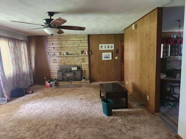 living room featuring a wood stove, wood walls, ceiling fan with notable chandelier, and dark colored carpet