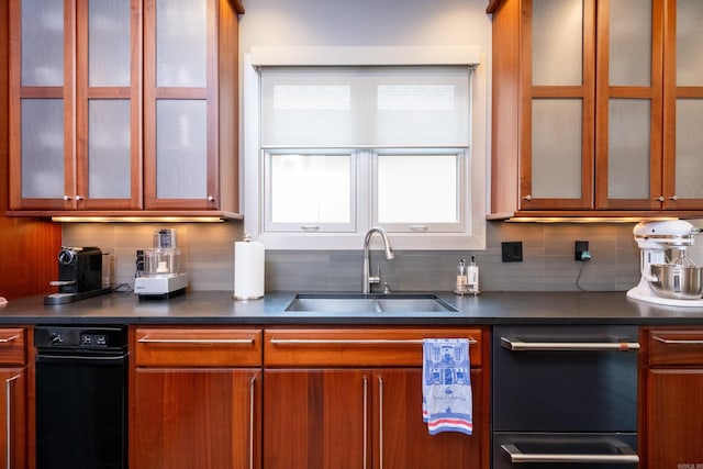 kitchen with a sink, tasteful backsplash, brown cabinetry, dark countertops, and glass insert cabinets