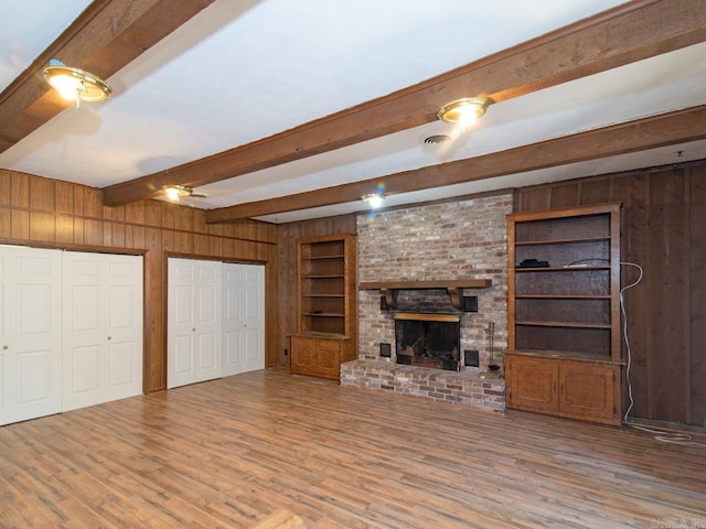 unfurnished living room featuring built in shelves, a brick fireplace, beamed ceiling, wood walls, and hardwood / wood-style floors