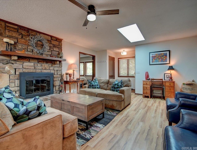 living room featuring a skylight, a textured ceiling, ceiling fan, light hardwood / wood-style flooring, and a stone fireplace