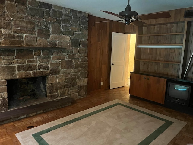 unfurnished living room with wood walls, a stone fireplace, ceiling fan, and dark parquet floors