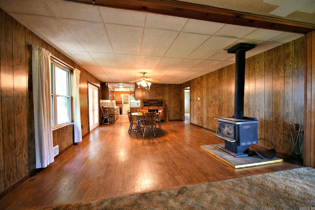 unfurnished dining area with a paneled ceiling, wooden walls, hardwood / wood-style floors, a wood stove, and ceiling fan