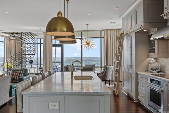 kitchen featuring an island with sink, oven, sink, a wall of windows, and dark wood-type flooring