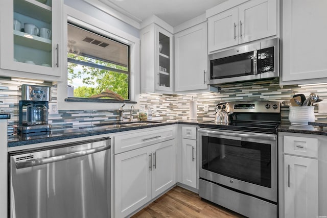 kitchen with stainless steel appliances, white cabinetry, decorative backsplash, dark stone counters, and glass insert cabinets