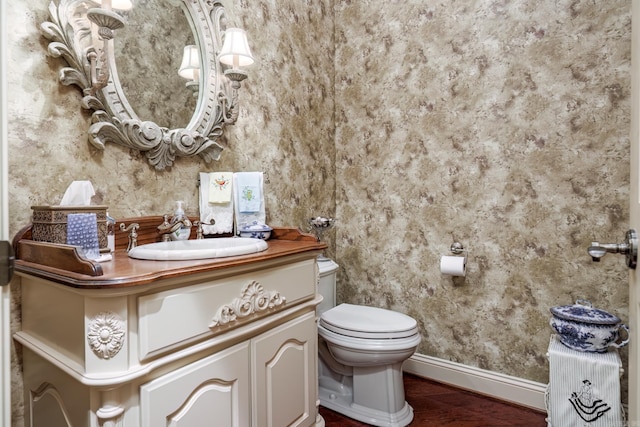 half bathroom featuring toilet, wood finished floors, wallpapered walls, baseboards, and vanity