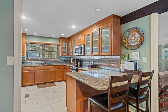 kitchen featuring glass insert cabinets, appliances with stainless steel finishes, brown cabinetry, a peninsula, and a kitchen breakfast bar