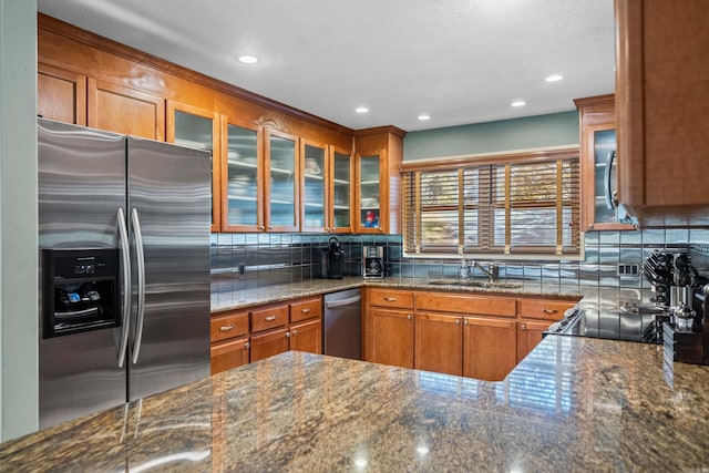 kitchen featuring appliances with stainless steel finishes, tasteful backsplash, brown cabinetry, dark stone countertops, and glass insert cabinets