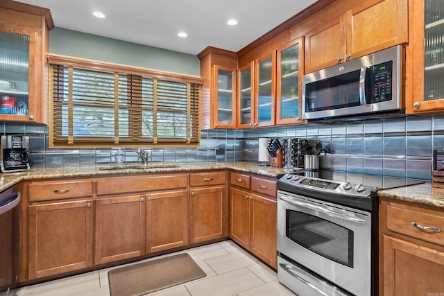 kitchen featuring light stone counters, brown cabinets, glass insert cabinets, stainless steel appliances, and a sink