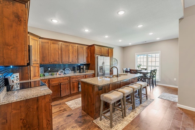 kitchen featuring a center island with sink, tasteful backsplash, light stone countertops, a kitchen bar, and stainless steel fridge with ice dispenser