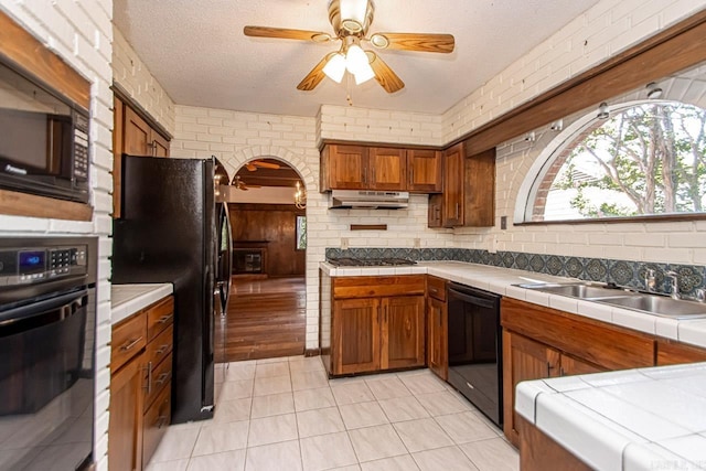 kitchen with brown cabinets, tile counters, a sink, brick wall, and black appliances