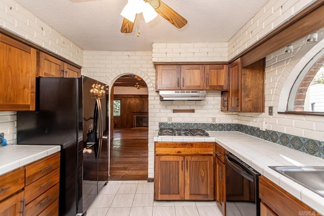 kitchen featuring arched walkways, under cabinet range hood, tile counters, black appliances, and brown cabinetry