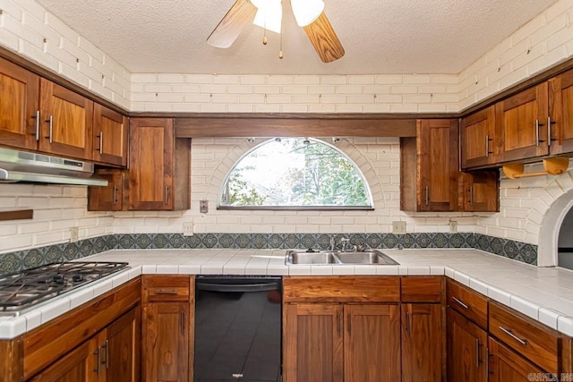 kitchen featuring tile counters, stainless steel gas stovetop, brown cabinetry, a sink, and under cabinet range hood