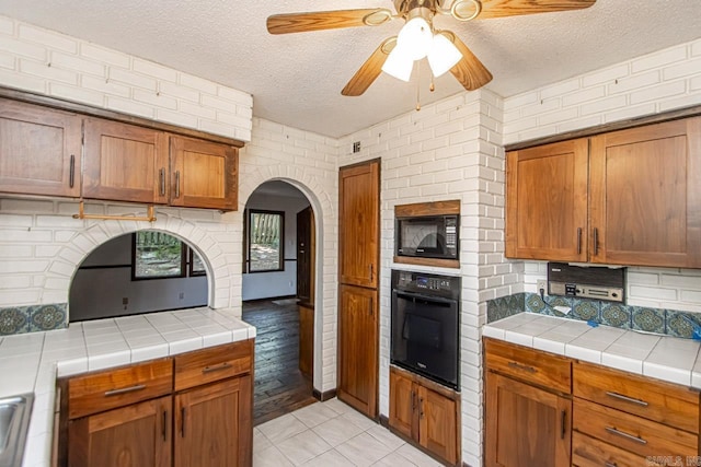 kitchen with brown cabinetry, a ceiling fan, tile countertops, a textured ceiling, and black appliances