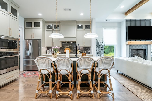 kitchen with appliances with stainless steel finishes, pendant lighting, tasteful backsplash, a kitchen island with sink, and light hardwood / wood-style floors