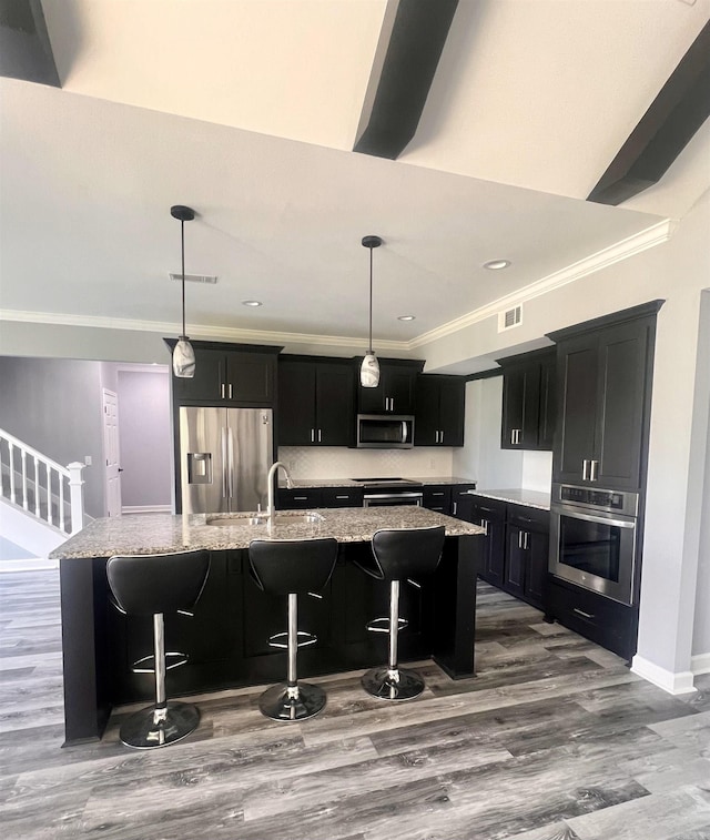 kitchen with appliances with stainless steel finishes, hardwood / wood-style flooring, light stone countertops, a breakfast bar area, and a center island with sink