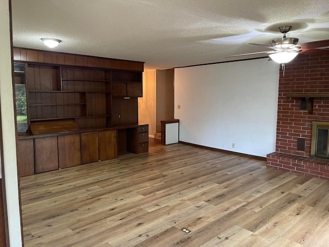 unfurnished living room featuring a brick fireplace, light hardwood / wood-style flooring, a textured ceiling, ceiling fan, and brick wall