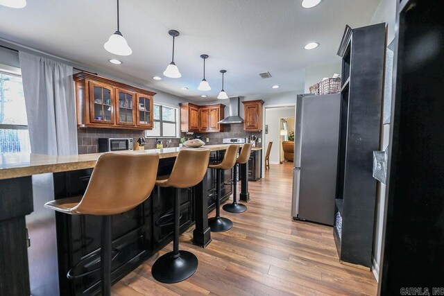 kitchen with backsplash, a kitchen bar, wall chimney range hood, stainless steel fridge, and hardwood / wood-style flooring