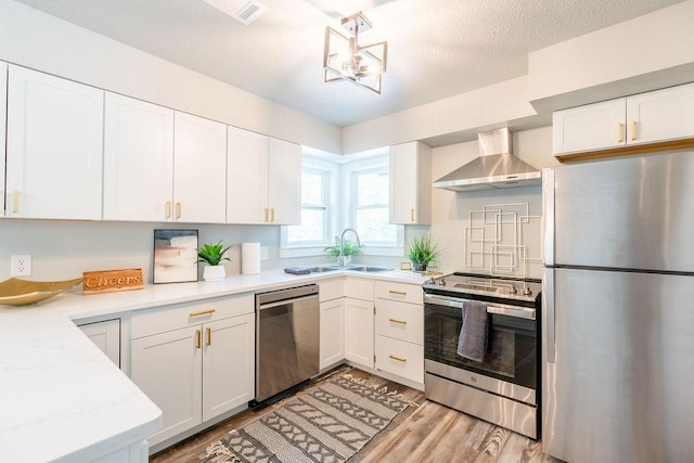 kitchen featuring light wood finished floors, stainless steel appliances, white cabinetry, a sink, and wall chimney range hood