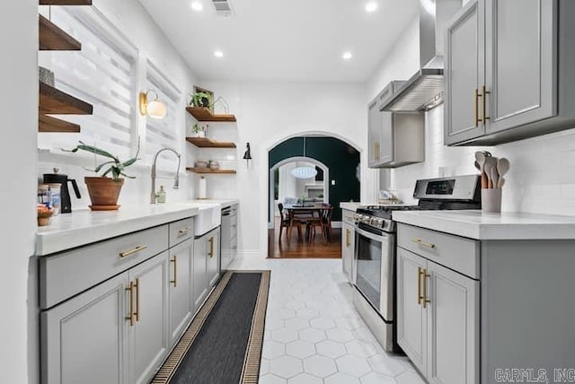 kitchen featuring backsplash, wall chimney exhaust hood, gray cabinetry, light tile patterned floors, and stainless steel stove