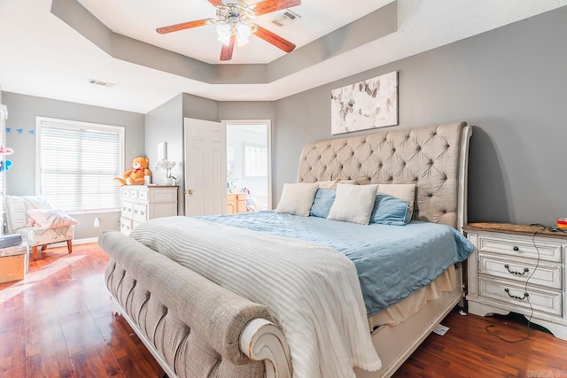 bedroom with baseboards, visible vents, a ceiling fan, dark wood-style floors, and a tray ceiling