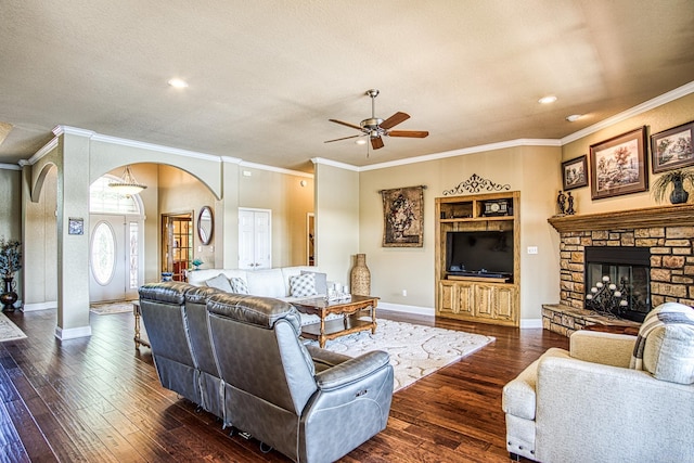 living room featuring a textured ceiling, a stone fireplace, dark wood-type flooring, ornamental molding, and ceiling fan