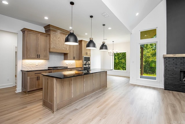 kitchen featuring a center island with sink, pendant lighting, a stone fireplace, sink, and light wood-type flooring