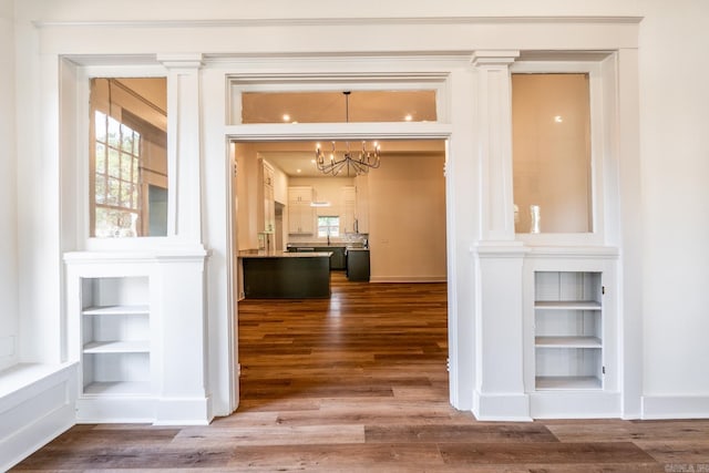 hallway with built in shelves, wood-type flooring, a chandelier, and ornate columns